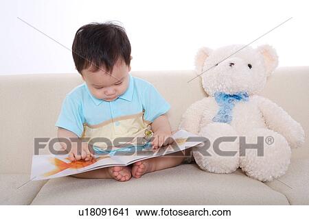Stock Image - Baby boy sitting on sofa looking at picture book. Fotosearch
