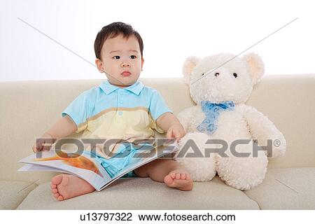 Stock Image - Baby boy sitting on sofa with picture book and teddy bear. Fotosearch
