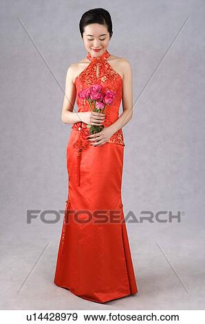 Stock Photo - Smiling young woman wearing a red embroidered cheongsam, holding pink roses, looking down. Fotosearch