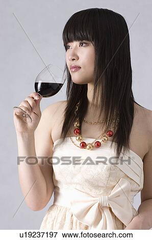 Stock Photo - Young woman wearing a light beige formal dress, holding a glass of wine to her mouth, side view. Fotosearch