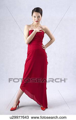 Stock Image - Young woman wearing a red evening gown and high heels, dressed up, hand on chest. Fotosearch
