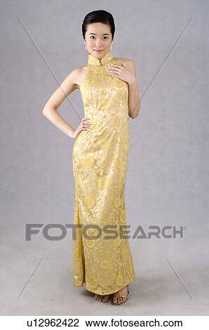 Stock Image - Young woman wearing a yellow embroidered cheongsam, hand on hip, smiling. Fotosearch
