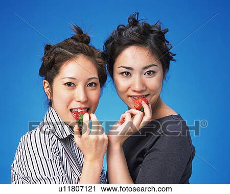 Stock Image - Two Young Women Eating Strawberry, Looking at Camera, Front View,. Fotosearch