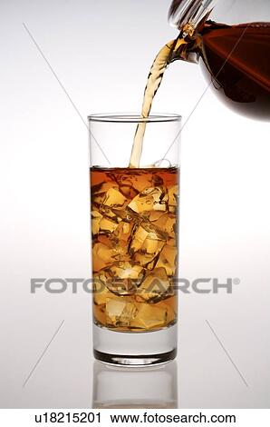 Stock Image - Black tea, pouring from a teapot into a glass filled with ice cubes. Fotosearch