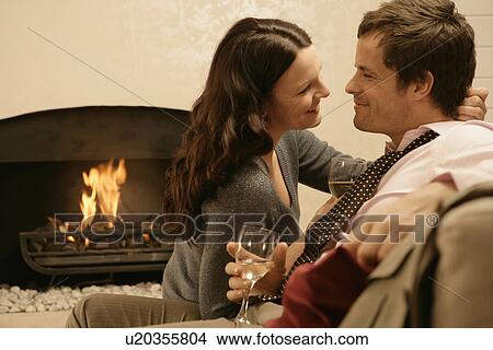 Picture - Mid adult couple drinking wine and hugging by fireplace. Fotosearch