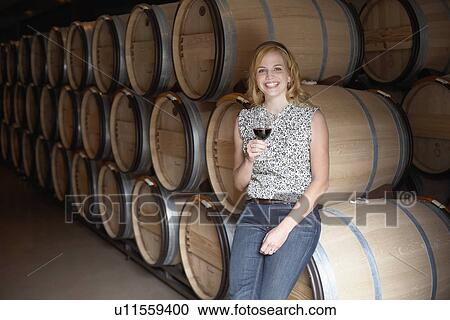 Stock Image - Woman drinking red wine by wine barrels (portrait). Fotosearch