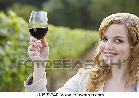 Stock Image - Young woman holding glass of red wine in vineyard (portrait). Fotosearch