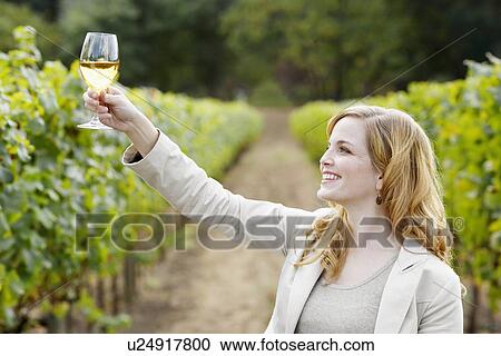 Stock Image - Young woman holding glass of white wine in vineyard. Fotosearch