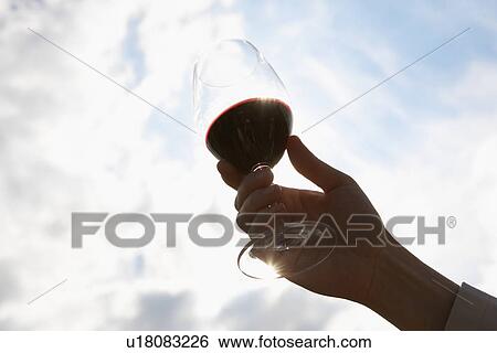 Stock Photograph - Person holding glass of red wine, sky in background (close-up, low angle view). Fotosearch