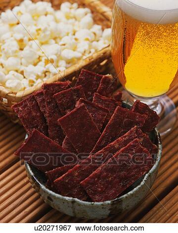 Stock Photo - Closed Up Image of a Little Bowl Filled With Beef Jerky, and a Wooden Basket Filled With Popcorn Next to a Glass of Beer, High Angle View. Fotosearch