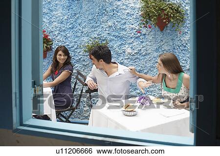 Stock Photograph - Man flirting with other woman in restaurant, girlfriend pulling him from shirt. Fotosearch