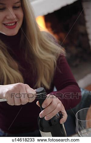 Stock Image - Young woman uncorking bottle of wine by the fireplace. Fotosearch