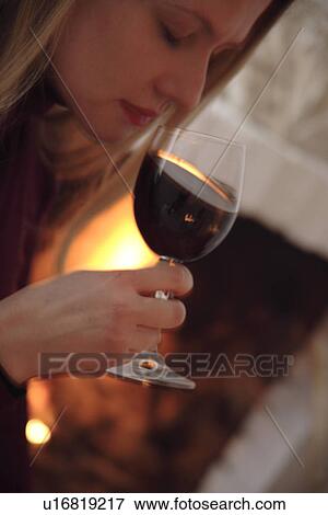 Stock Photo - Young woman tasting glass of red wine by the fireplace, side view. Fotosearch