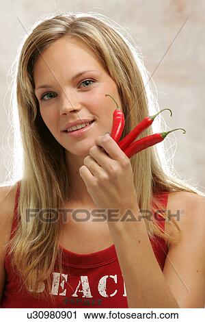 Stock Image - Young woman with long blond hair holding three chili peppers, portrait. Fotosearch