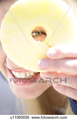 Stock Photography - Portrait of young woman holding pineapple slice in front of one eye, smiling. Fotosearch