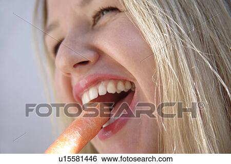 Stock Photograph - Young blond woman biting in fresh carrot, side view, close up. Fotosearch