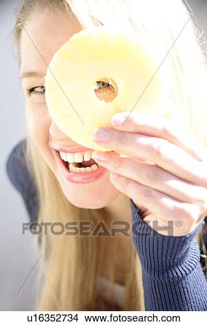 Picture - Portrait of young woman holding pineapple slice in front of one eye, smiling. Fotosearch