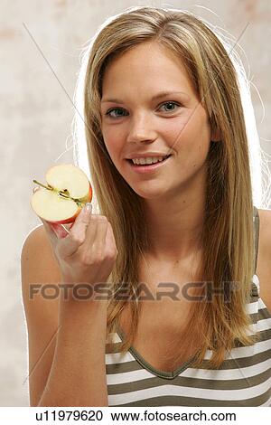 Stock Image - Young woman with long blond hair eating an apple, portrait. Fotosearch