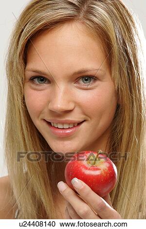 Stock Image - Young woman with long blond hair eating an apple, portrait. Fotosearch