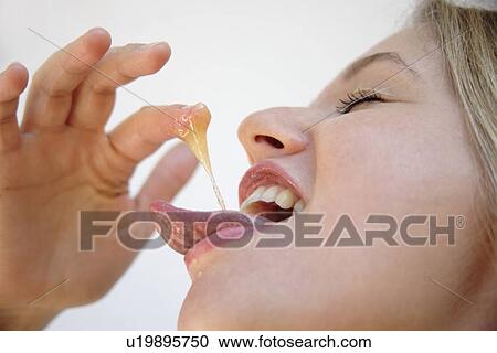 Stock Image - Young blond woman licking honey off her finger, side view, close up. Fotosearch