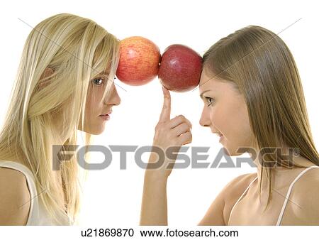 Stock Image - Side profile of two young women balancing apples with their foreheads. Fotosearch