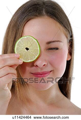 Picture - Close-up of a young woman holding a slice of lime in front of her eye. Fotosearch