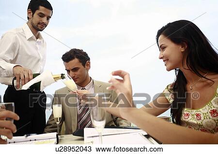 Stock Image - Man and woman at table with others being served champagne. Fotosearch