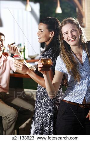 Stock Image - Two women in a bar with man in background. Fotosearch