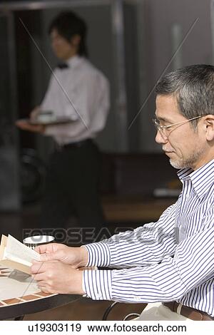 Stock Photo - A Mature Adult Man Reading a Book at a Cafe, Side View, Differential Focus. Fotosearch