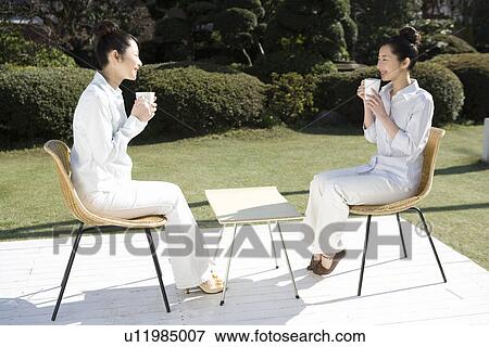 Stock Photo - Twin sisters sitting on chairs and holding cups in garden, smiling, side view. Fotosearch