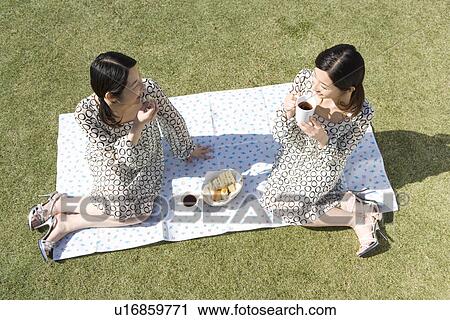 Stock Image - Twin sisters sitting on sheet in garden, tea time, high angle view. Fotosearch