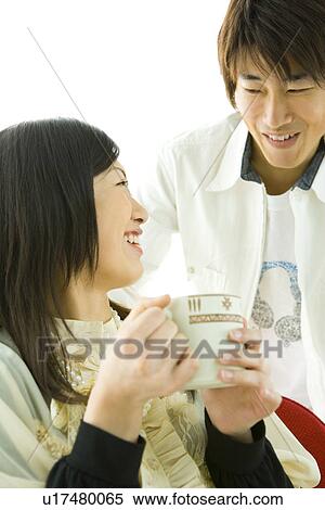 Stock Photography - Young man and woman smiling and looking at each other, woman holding a mug, front view, side view, white background. Fotosearch