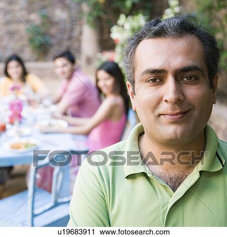 Stock Image - Portrait of a mid adult man smiling with his friends sitting at a dining table in the background. Fotosearch