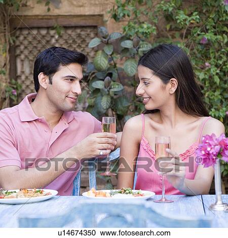 Stock Image - Young couple sitting at a dining table and holding champagne flutes. Fotosearch