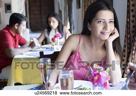 Stock Photo - Young woman sitting in a restaurant with a mid adult couple having food in the background. Fotosearch