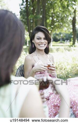 Picture - Two young women holding cups of tea and sitting at a table. Fotosearch