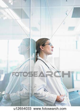 Stock Image - Concerned doctor leaning on glass wall in hospital lobby.. Fotosearch