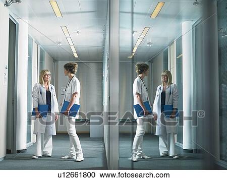 Stock Image - Two female doctors in discussion in a lobby of a hospital. Reflection on glass wall.. Fotosearch