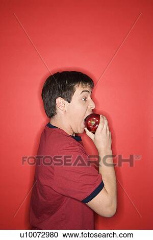 Stock Image - Portrait of Caucasian teen boy biting an apple standing against red background.. Fotosearch