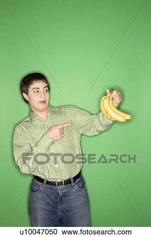 Stock Image - Portrait of Caucasian teen boy holding and pointing at bananas standing against green background.. Fotosearch