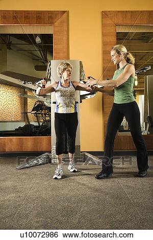 Prime adult Caucasian female helping mature adult Caucasian female use exercise equipment at gym. View Large Photo Image Stock Photograph - Prime adult Caucasian female helping mature adult Caucasian female use exercise equipment at gym.. Fotosearch