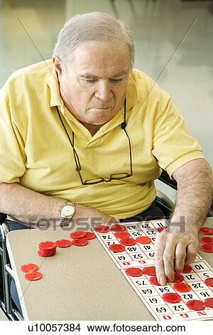 Picture - Elderly Caucasian man sitting in wheelchair playing bingo at retirement community center.. Fotosearch