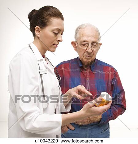 Stock Photo - Mid-adult Caucasian female doctor pointing at prescription bottle as elderly Caucasian male looks at bottle.. Fotosearch