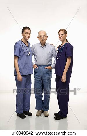 Stock Image - Two Caucasian females wearing scrubs standing with elderly Caucasian male.. Fotosearch