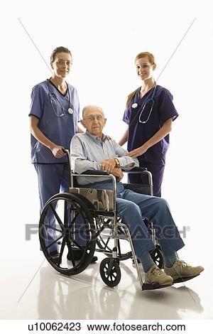 Stock Image - Two Caucasian females wearing scrubs with elderly Caucasian male in wheelchair.. Fotosearch
