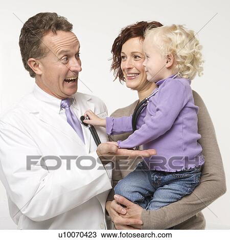 Stock Image - Caucasian female toddler holding stethoscope up to middle-aged male doctor smiling.. Fotosearch