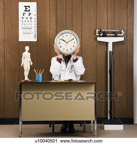 Stock Image - Caucasian male doctor sitting at desk holding clock over face.. Fotosearch