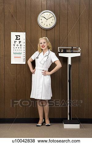 Stock Photo - Caucasian mid-adult female nurse with hands on hips smiling at viewer in retro doctor's office.. Fotosearch