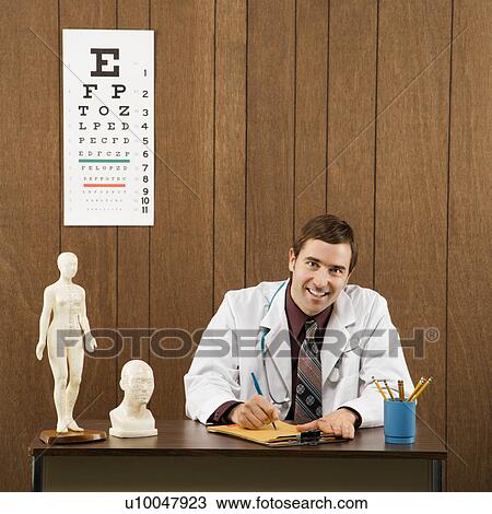 Stock Image - Mid-adult Caucasian male doctor sitting at desk writing and looking at viewer.. Fotosearch