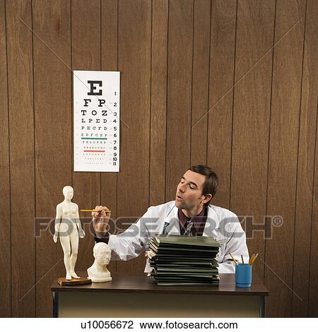 Stock Image - Mid-adult Caucasian male doctor sitting at desk pointing to figurine.. Fotosearch